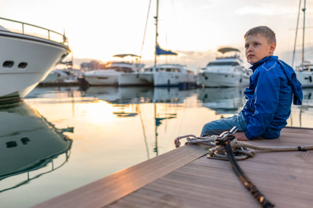 istock A boy in a blue jacket sits on a pier with yachts during sunset. 1202636672