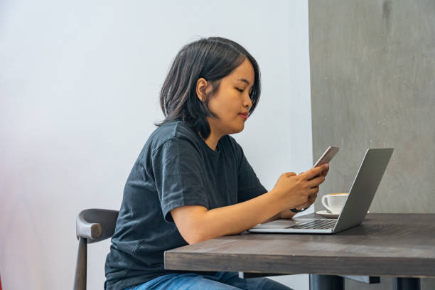 Asian woman freelancer using smartphone while working on laptop stock photo