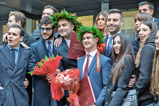 Rome, Italy - December 19, 2019: A group of young people including two males in the centre weraing laurel wreathes and holding books and bouquets, posing for a photographer on the left  (out of frame)  before a University faculty in Rome, Italy, without direction from me.