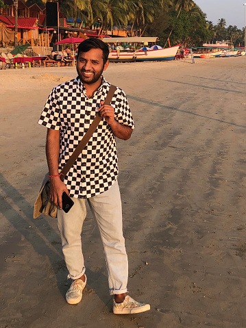Stock photo showing young Indian man walking by sea on wet sand on beach holiday on wet sand with shoulder bag / messenger bag on shoulder holding mobile phone, wearing black and white checked shirt, blue denim trousers, good looking Hindu man laughing smiling, walking on beach with shoes on Kerala Goa seaside, India