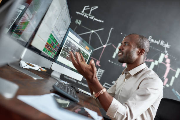 me siento mal cuando llegas tarde. retrato de hombre de negocios africano con estilo, comerciante sentado por el escritorio frente a múltiples monitores. se está enojando mientras mira los gráficos actuales - stock exchange stock market stock certificate wall street fotografías e imágenes de stock