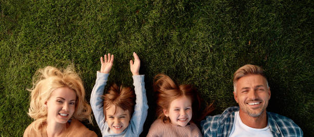 Keep calm and love your family. Happy family of four lying on green grass Top view of smiling parents, little girl and boy having fun while lying on a grass. Children, family and nature concept. Horizontal shot web banner photos stock pictures, royalty-free photos & images