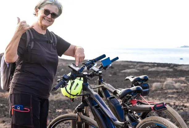 Photo of A senior woman athlete indicates the electric bicycle device that allows her to travel with little effort. Excursion on the cliff, the sea in background