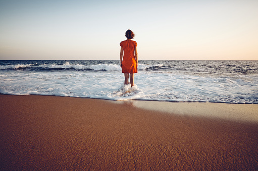 Rear view of a woman in orange dress standing still in the sea at sunset, color toning applied.