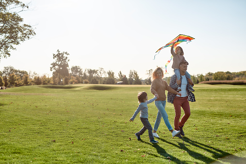 Full-length portrait of cheerful parents with two kids running with kite in the park on a sunny day. Family, kids and nature concept. Horizontal shot.