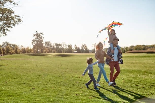 Children make life important. Happy family playing a kite. Outdoor family weekend Full-length portrait of cheerful parents with two kids running with kite in the park on a sunny day. Family, kids and nature concept. Horizontal shot. public park stock pictures, royalty-free photos & images