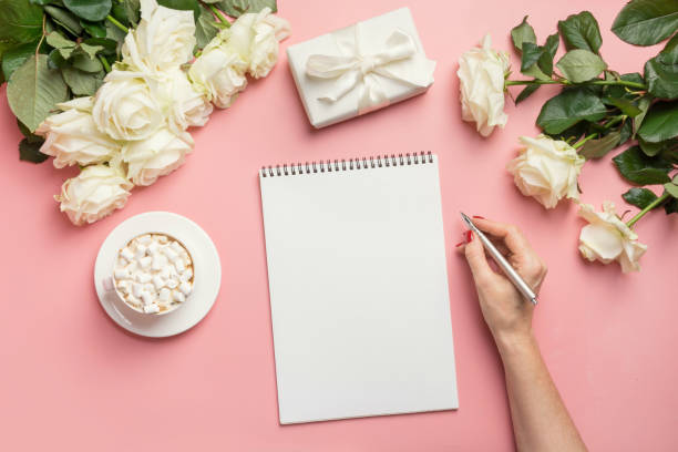 istock Female hands writing in notebook and bouquet white roses on pink. Top view with copy space. 1202695544