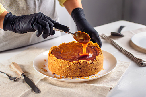 Young woman in gloves decorating cheesecake with caramel in the kitchen