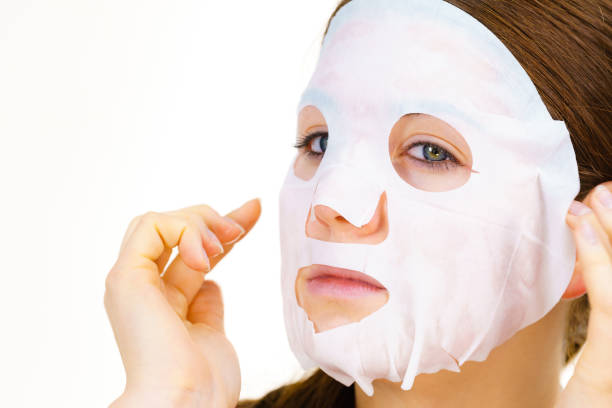 Woman applying sheet mask on her face, on white. Girl taking care of skin complexion. Beauty treatment. Skincare.