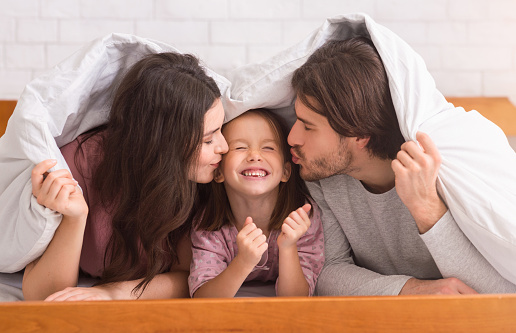 Family Protection. Loving Mom And Dad Kissing Cheeks Of Their Adorable Little Daughter And Hiding Under Blanket In Bed, Panorama
