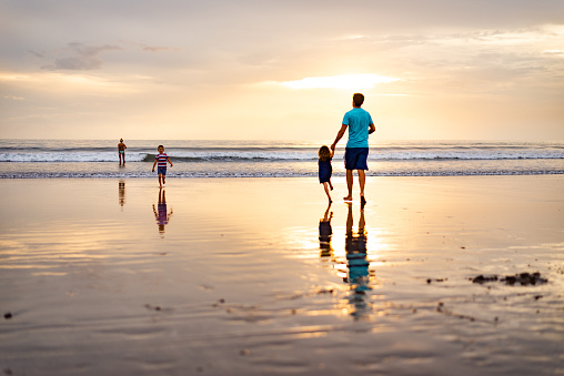 Rear view of parents and their little kids, a boy and a girl, having fun while running towards the sea on the beach at dusk. Copy space.