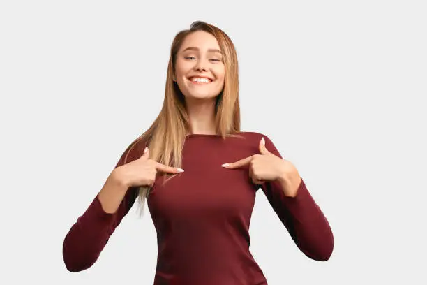 Girl points at herself with index fingers and shows off successes and achievements. Beautiful young woman with long hair, wears Burgundy-colored jacket sleeves isolated on white background in Studio
