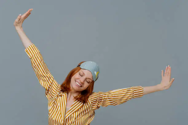 Perfect rest and sleeping concept. Cheerful young ginger European woman spreads arms and stretches after awakening, had sweet dreams, wears striped pajama and sleepmask, isolated on grey wall