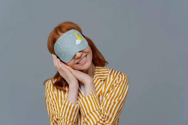 Photo of Just relaxing or time for nap. Smiling gentle woman with eye mask, tilts head on pressed palms, wears striped pajama, poses over grey wall, copy space area for your text feels comfortable and relaxed