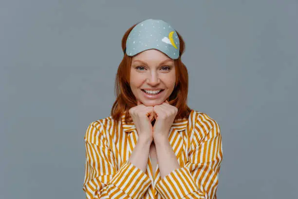 Headshot of attractive woman keeps hands under chin, has ginger hair, pleasant toothy smile, wears yellow nightwear and sleepmask, looks directly at camera, isolated over grey background. Wake up