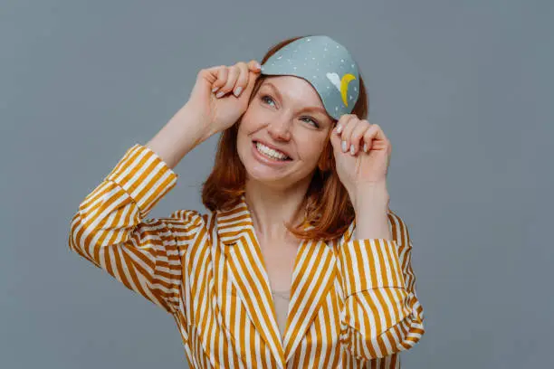 Dreamy redhaired European woman looks gladfully aside, smiles broadly, wears casual domestic clothes and sleepmask, isolated over grey background, imagines something pleasant. Good morning concept