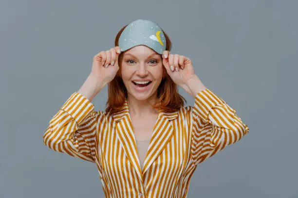 Ginger female model wears sleepmask and striped pajamas, awakes in good mood, realizes today is weekend, can stay in bed for long time, smiles positively, isolated on grey background. Sleeping