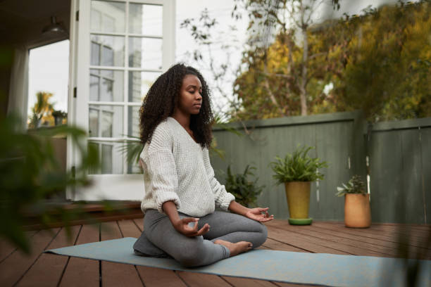 Young woman sitting in the lotus pose outside on her patio Young African American woman sitting on exercise mat outside on her patio and meditating in the lotus pose during a yoga session meditating stock pictures, royalty-free photos & images