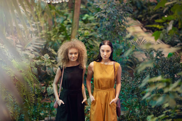 Portrait of two beautiful young women standing in stylish dresses and looking at camera with green plants and trees in the background