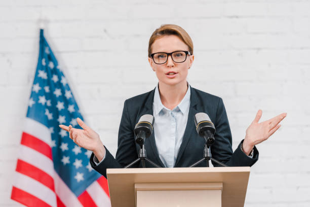 istock beautiful speaker in glasses gesturing while talking near microphones 1202730396