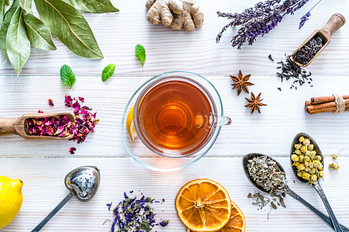 Herbal tea cup surrounded by a variety dried tea leaves and flowers in spoons shot from above on white table. The composition includes dried part of plants like hibiscus, calendula, ginger, rose petals, chamomile, bay leaves, cinnamon sticks, star anise, a tea strainer and dried orange slices among others. High resolution 42Mp studio digital capture taken with Sony A7rii and Sony FE 90mm f2.8 macro G OSS lens