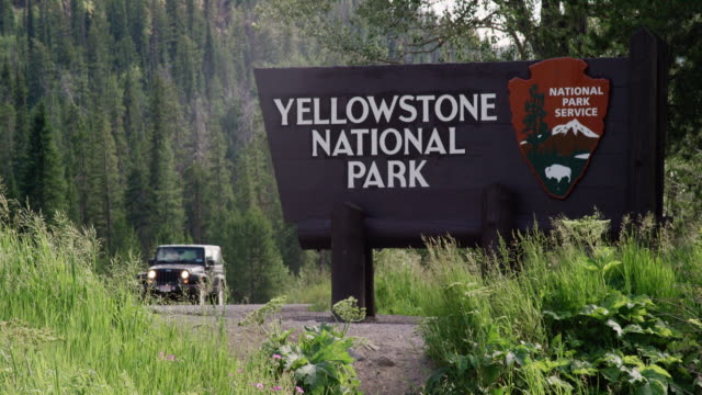 A Jeep and Motorcycles Drive by the Yellowstone National Park Welcome Sign Surrounded by Forest and the Rocky Mountains