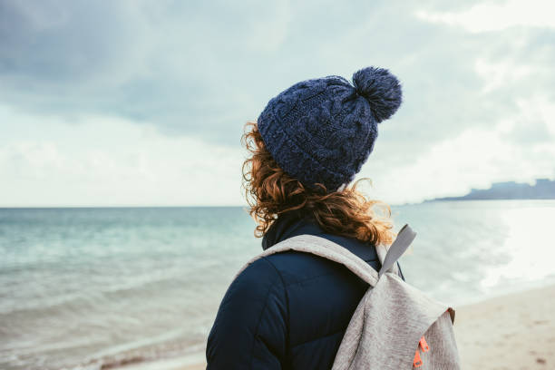 View from behind young woman dressed in knitted hat standing looking at winter sea.