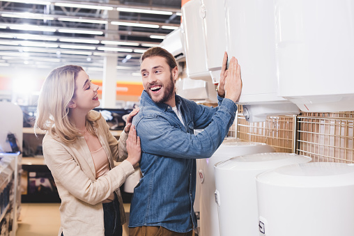 smiling boyfriend touching boiler and looking at girlfriend in home appliance store