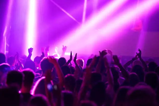 Photo of Blurred image of silhouette of raised arms, crowd of people in the front of bright stage lights at concert