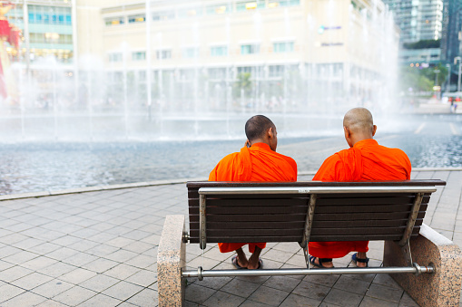 Kuala Lumpur / Malaysia - 02.14.2018: Two Buddhist monks relax on a bench in the park, watching the fountain.