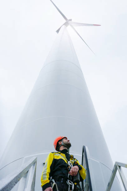 Young rope access technician standing under wind turbine and looking up stock photo