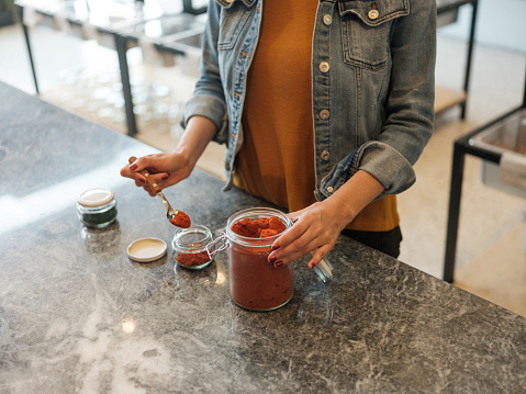 A young latin woman filling up a glass container by using a spoon at the zero waste shop.