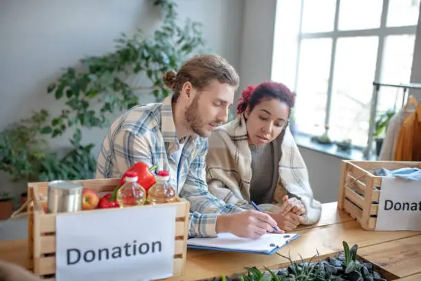 Social issues. Young bearded man with notebook and girl with colored hair sitting at a table with charity food boxes, solving social issues.