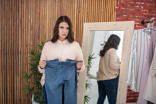 Overweight. Young woman in a beige blouse holding new jeans and feeling stressed