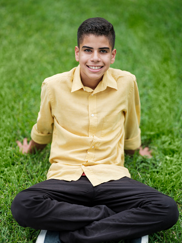 A latin teenage boy sitting on the grass, leaning back and smiling at the camera.