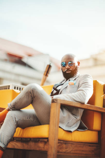 An afro man in a plaid suit A vertical shot an elegant mature bearded bald African man in round bluish sunglasses and a checkered suit sitting on a yellow outdoor sofa and looking at the camera; a black guy in a plaid suit luxury eyewear stock pictures, royalty-free photos & images