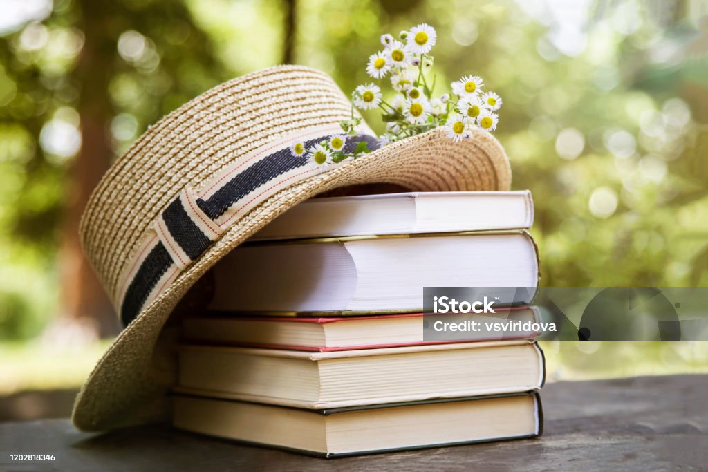 Rural summer still life in the park among trees. A stack of books on an old wooden table next to a straw hat of a canoe and a bouquet of wild flowers. Book Stock Photo