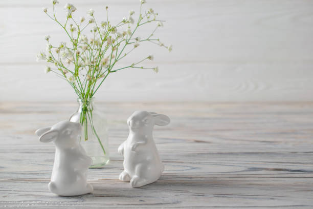 Easter decoration. Ceramic white bunnies on white wooden table with fresh white spring flowers.