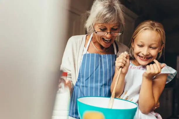 Happy grandmother and kid mixing a cake batter at home. Happy little girl stirring batter in a bowl with her granny standing by her side.