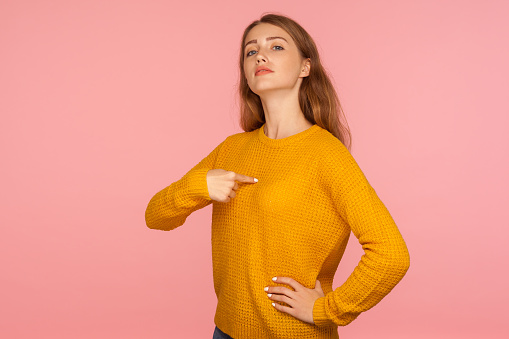 This is me! Portrait of attractive haughty ginger girl in sweater pointing at herself and looking at camera with arrogance, proud and confident in success. studio shot isolated on pink background