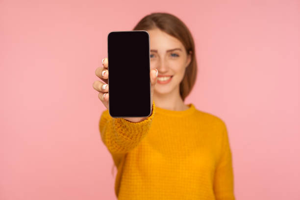 portrait of fascinating happy ginger girl in sweater showing cellphone to camera and smiling, pleased satisfied with device and application - photography human hand portrait women imagens e fotografias de stock