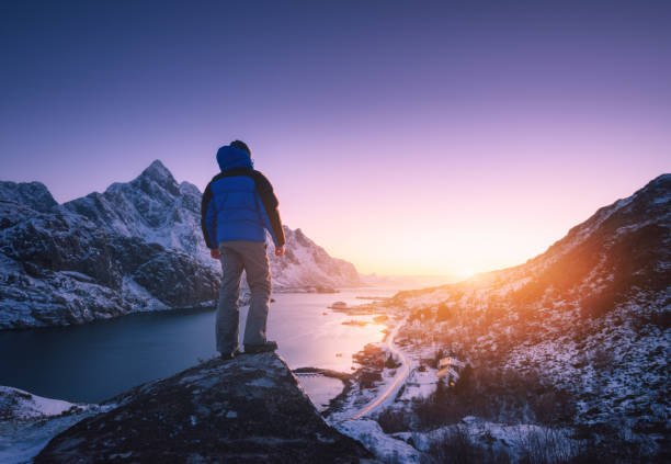 Man is standing on the mountain peak against snowy mountains, fjord at colorful sunset in winter. Young man on the stone, sea coast and rocks, blue sky in Lofoten Islands, Norway. Travel and hiking Man is standing on the mountain peak against snowy mountains, fjord at colorful sunset in winter. Young man on the stone, sea coast and rocks, blue sky in Lofoten Islands, Norway. Travel and hiking northern norway stock pictures, royalty-free photos & images