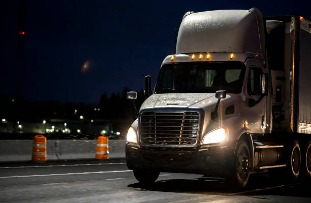 Big rig white day cab semi truck transporting cargo in semi truck driving on the wet rainy road in dark night time Big rig day cab heavy-duty diesel semi truck with roof spoiler transporting commercial cargo in semi trailer running on the wet raining highway road at dark night time with turned on headlight truck in rain stock pictures, royalty-free photos & images