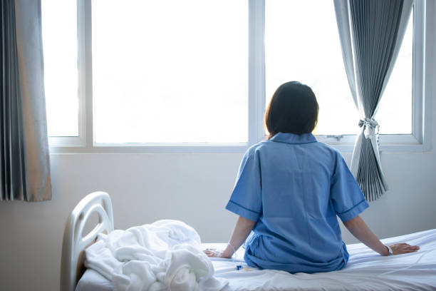 back view of patient woman sitting on bed in hospital ward, looking away at window and hope everything will be better. - depression women sadness window imagens e fotografias de stock