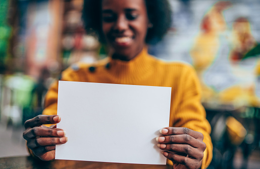 Young beautiful african-american woman holding in hands white empty blank paper page with copy space.