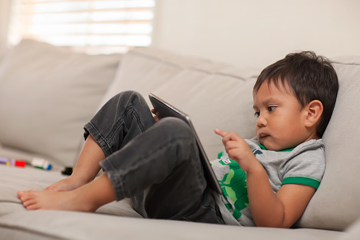 A young kid, 3 years old who is using his little finger on the touchscreen of his big tablet, while he watches  the internet on a comfy couch.