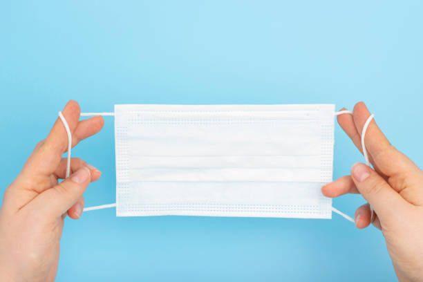 istock Female hands holding white mask on blue background, top view 1202870688