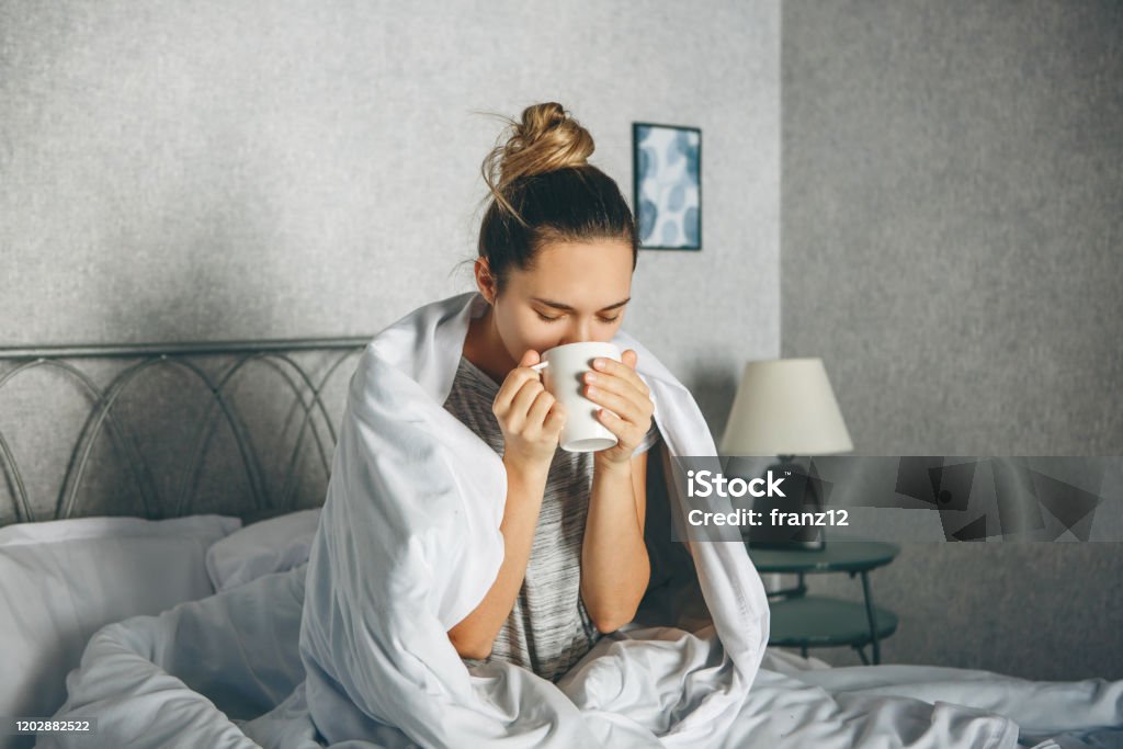 Girl drinks a drink from a mug Girl drinks a drink from a mug in bed wrapped in a blanket. She is enjoying or she is not awake or sick. Illness Stock Photo