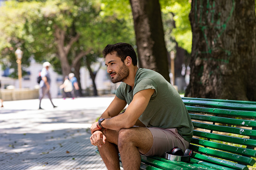 A Latino man is sitting on a park bench by himself in Buenos Aires.