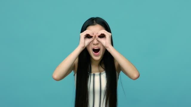 Surprised and curious asian woman with long black hair looking through fingers in binoculars gesture